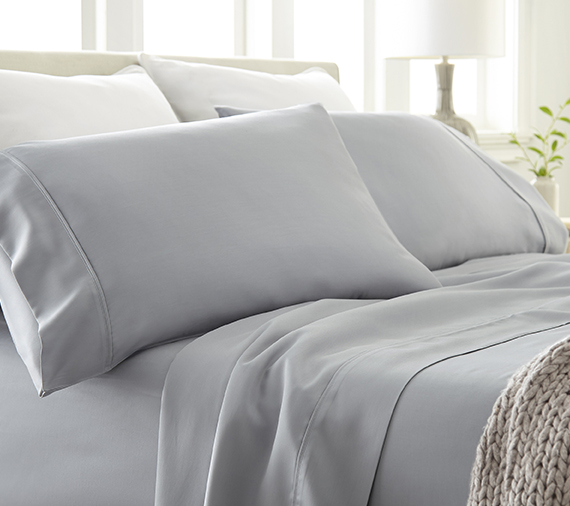 Bed with multiple pillow and small plant on stand in room with windows open and sun shining